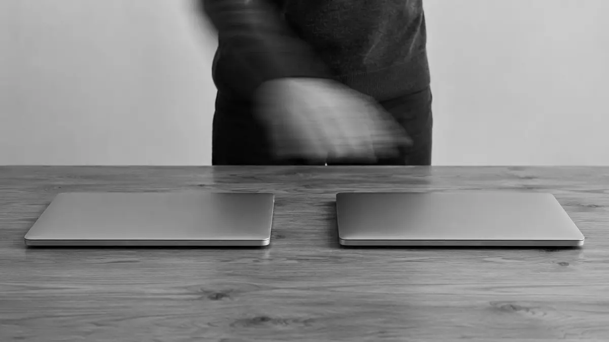 A blurred hand moves above two closed laptops placed side by side on a wooden table.