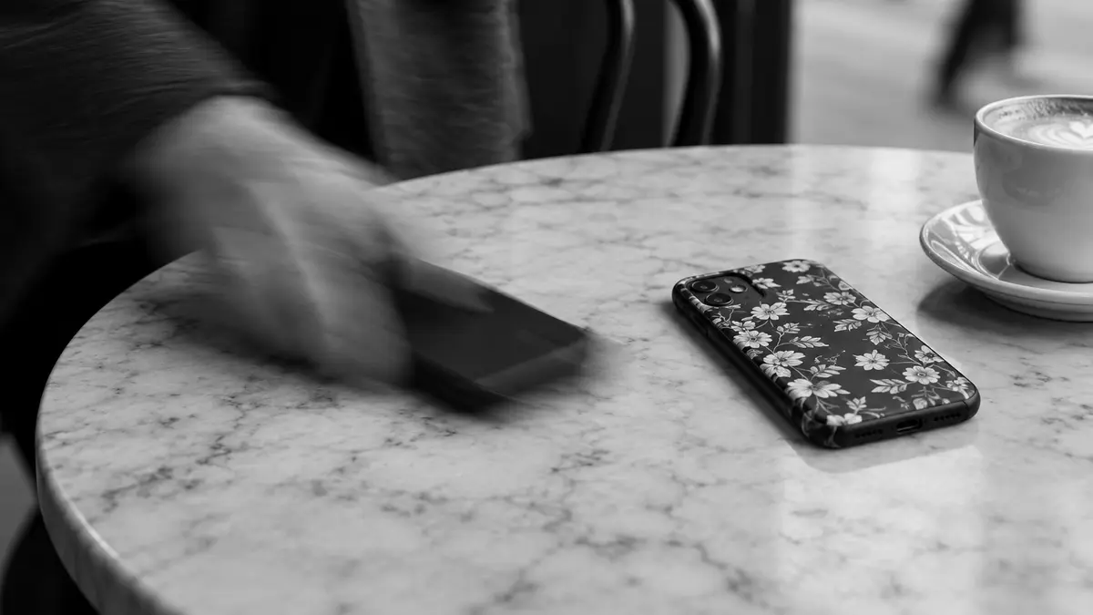 Une main floue déplace un téléphone noir près d’un téléphone à coque fleurie et d’une tasse de café.